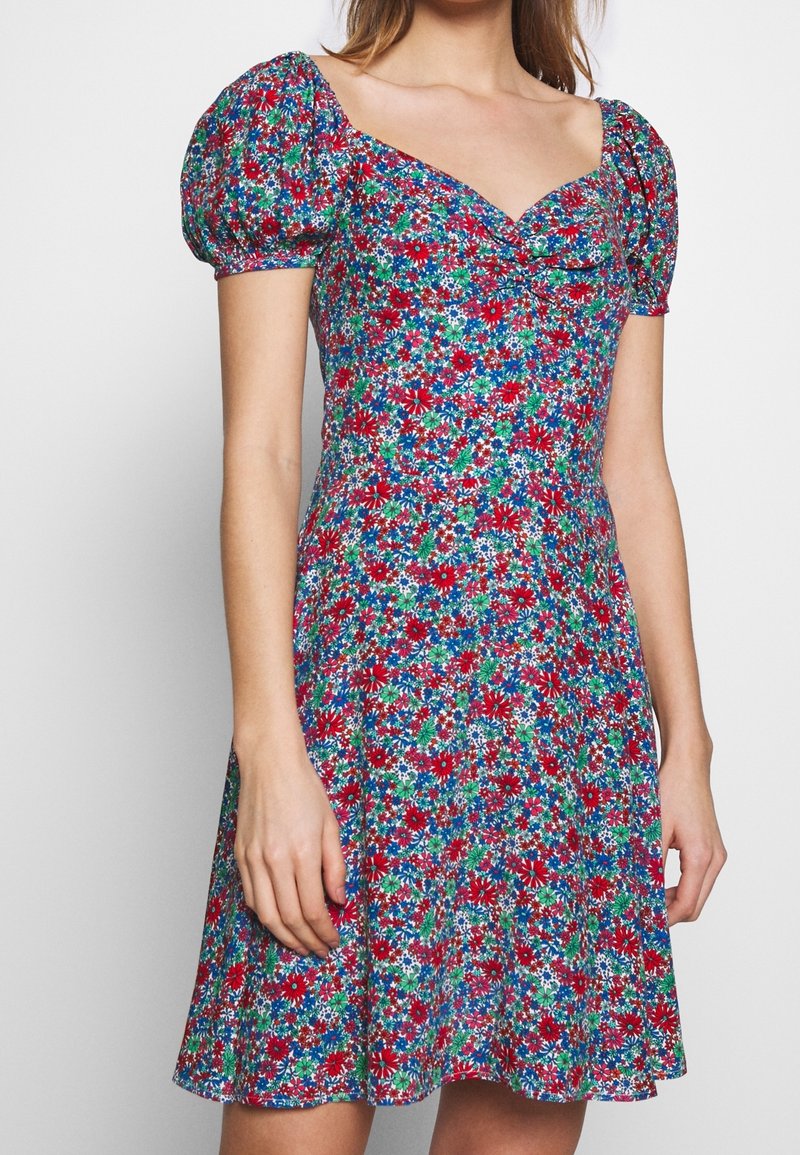 Femme portant une robe courte à manches bouffantes avec un motif floral rouge, bleu, vert et blanc et un décolleté en cœur.