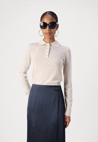 Beige langslavige polo trui met knopenpaneel en geribde manchetten, gecombineerd met een vloeiende donkerblauwe rok. Zwarte oversized zonnebril gedragen.