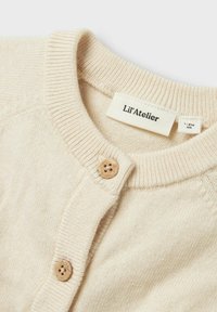 Beige Strickjacke mit Rundhalsausschnitt, zwei braunen Knöpfen, gerippten Bündchen und einem Etikett mit der Aufschrift "Lil'Atelier" im Inneren.