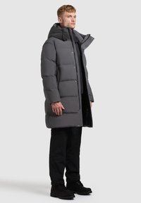 Szürke párnázott kabát, cipzáros elülső részes és kapucnis. Nagy zsebekkel és steppelt textúrával rendelkezik. Fekete nadrággal és csizmával van párosítva.