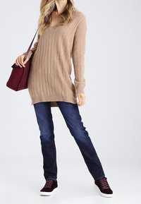 Femme portant un pull beige à col en V, un jean bleu foncé, des baskets bordeaux et un sac à bandoulière assorti bordeaux.