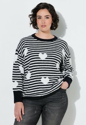 Vrouw met kort krullend haar die een zwart-wit gestreepte trui met hartpatronen en donkere spijkerbroek met strassdetails draagt.
