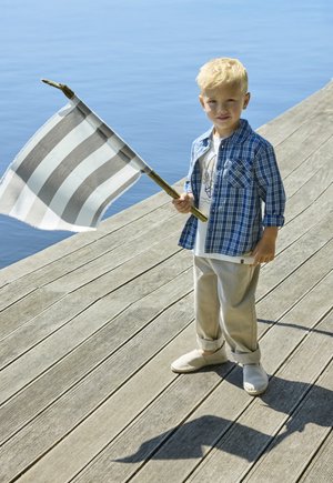 Enfant tenant un drapeau rayé fixé sur un poteau en bois, portant une chemise à carreaux bleue, un t-shirt blanc avec un motif, un pantalon beige et des chaussures claires.
