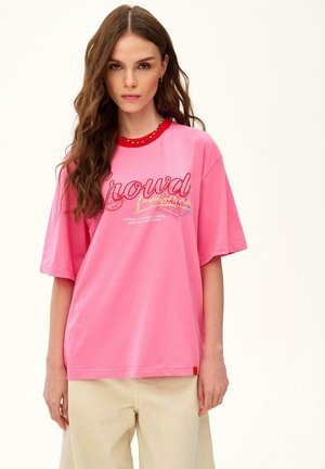 Młoda kobieta z długimi brązowymi włosami, ubrana w różowy oversize T-shirt z czerwonym kołnierzykiem i beżowe spodnie, stojąca na jednolitym tle.