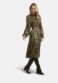 Donna che indossa un vestito midi a maniche lunghe con cintura e stampa leopardata, abbinato a stivali neri alti al ginocchio con tacchi, posando con un piede incrociato sull'altro.