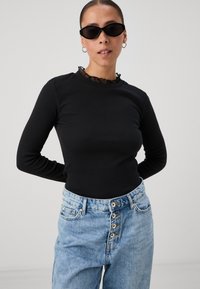 Black long-sleeve top with a ruffled neckline, paired with light blue high-waisted jeans featuring button details and a relaxed fit.