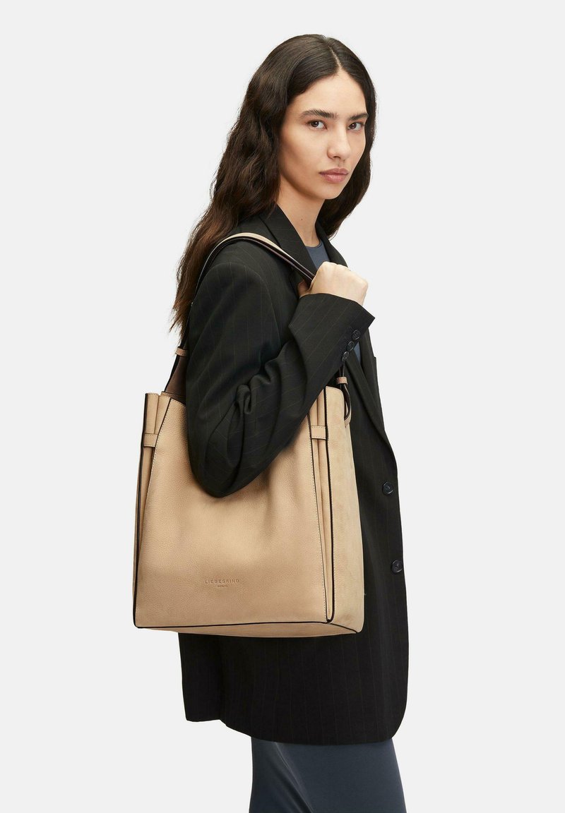 Frau mit langen dunklen Haaren, die ein schwarzes Nadelstreifen-Blazer trägt, trägt eine große beigefarbene Ledertasche über der Schulter.