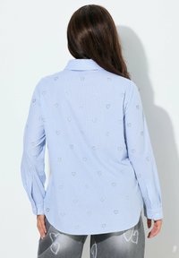 Frau trägt hellblau-weiß gestreiftes Hemd mit kleinen Herzmustern, kombiniert mit grauer Hose mit großen weißen Herzmotiven.