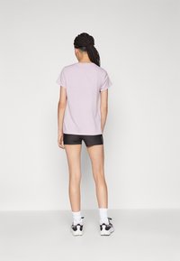 Fialové tričko s krátkým rukávem a kulatým výstřihem, v kombinaci s černými kraťasy. Látka vypadá měkce; detaily zahrnují malé logo na kraťasech.