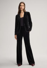 Donna in piedi che indossa un completo in velluto nero con blazer a un bottone e pantaloni a gamba larga, abbinati a scarpe a punta rosso scuro.