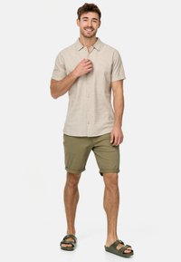 Camisa corta de manga en color beige claro con diseño de botones, combinada con pantalones cortos enrollados en color verde olivo y sandalias marrones. Vestimenta sencilla y casual.
