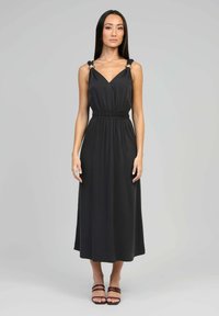 Femme se tenant dans une robe midi noire sans manches avec des détails en anneau métallique sur les bretelles et des sandales à talons marron, contre un fond gris.