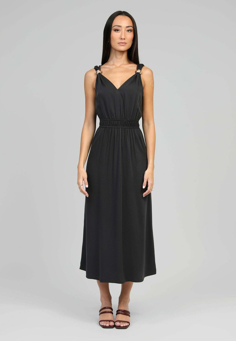 Femme se tenant dans une robe midi noire sans manches avec des détails en anneau métallique sur les bretelles et des sandales à talons marron, contre un fond gris.