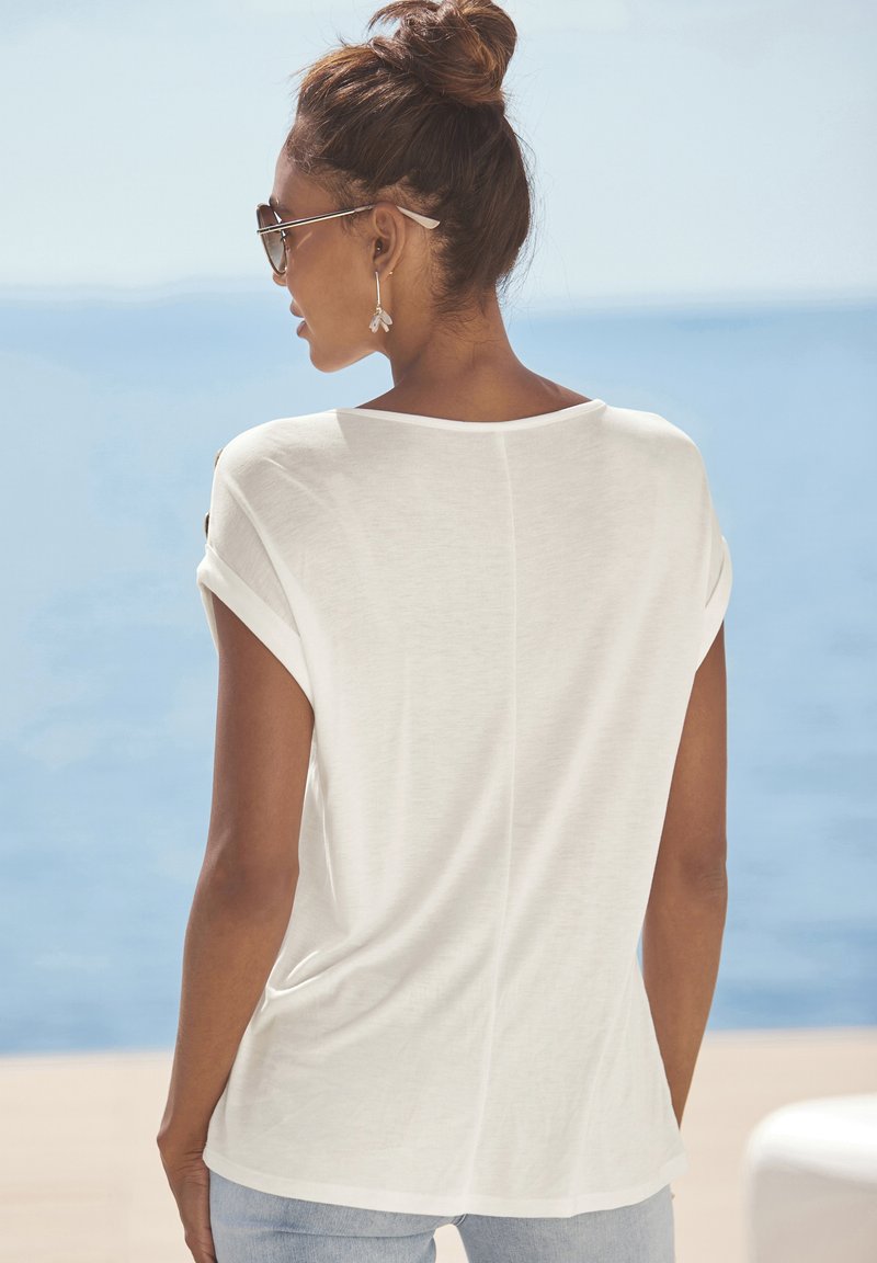 Witte korte mouwen top met een losse pasvorm, zachte textuur en opgerolde mouwen. Opvallende achterkantnaad, gecombineerd met lichtblauwe jeans.