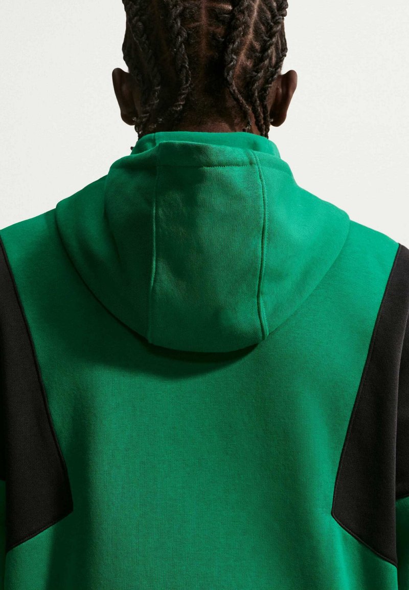 Persona con capelli intrecciati che indossa una felpa verde con pannelli neri, mostrata di spalle su uno sfondo chiaro uniforme.