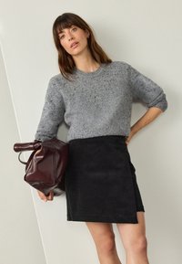 Gray knitted sweater with short sleeves, paired with a black corduroy skirt. Model holds a burgundy leather bag.