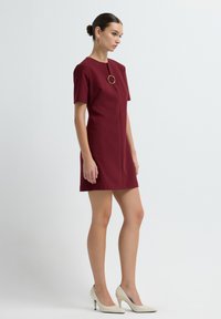 Femme se tenant dans une robe bordeaux à manches courtes avec un anneau doré décoratif au niveau du col, portant des chaussures à talons hauts blanches.