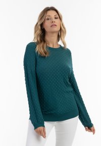Maglione lungo a maniche lunghe di colore verde bluastro con un motivo trapuntato, polsini e orlo a coste. Abbinato a pantaloni bianchi, con scollatura rotonda.
