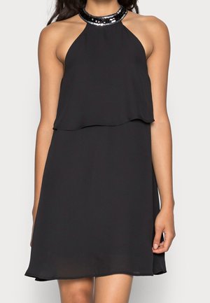 Femme portant une robe noire sans manches à col halter avec un corsage superposé et un col brillant en sequins noirs, debout les bras détendus le long du corps.