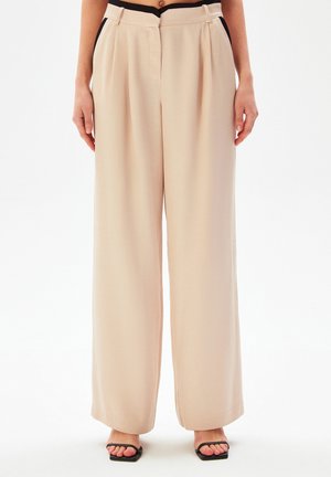 Cremfarbene, plissierte, weite Hosen aus glattem Stoff mit hoher Taille und seitlichen Taschen, kombiniert mit flachen, schwarzen Sandalen.