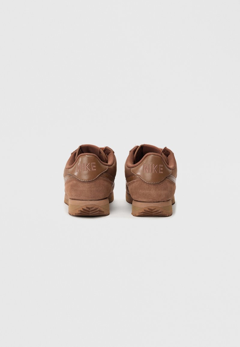 Brown suede sneakers with a textured finish, featuring a rubber sole and embossed "NIKE" logo on the heel. Classic athletic design.