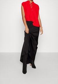 Rød plisseret bluse med korte ærmer, parret med en sort asymmetrisk nederdel og strukturerede sorte knæhøje støvler.