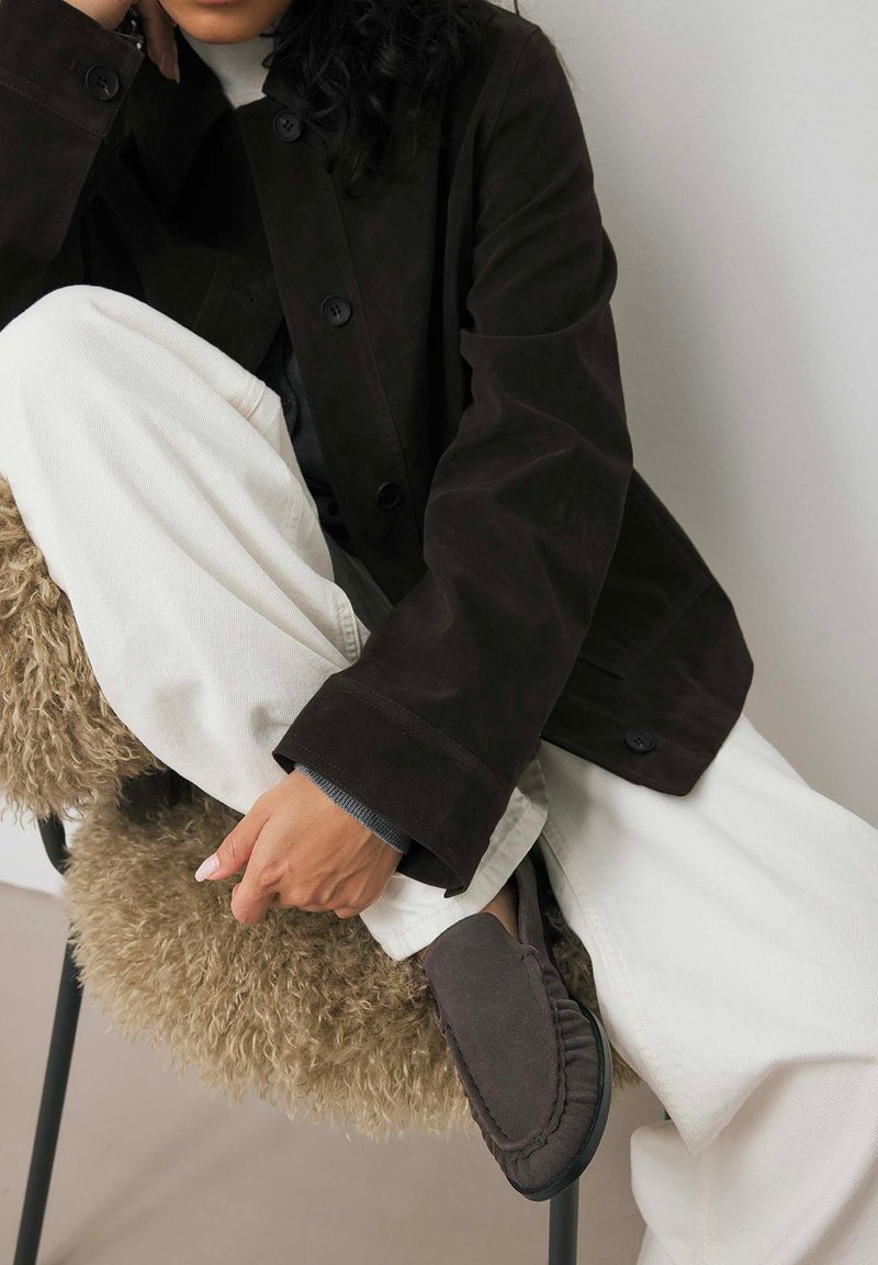 Tumepruun sulejakk nööpidega, kombineeritud kreemjatest laiematest püksidest ja hallidest mokassinsidest, istudes tekstuuriga toolil.