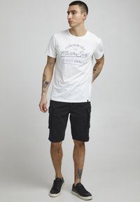 T-shirt blanc avec texte graphique bleu, associé à un short cargo noir avec poches latérales. Des baskets noires complètent la tenue.