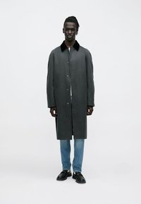 Harmaa polvipituinen takki, jossa musta kaulus, nepparikiinnitys ja sivutaskut, päällä vaaleansiniset farkut ja mustat kengät.