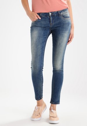 Jeans ajustados de mezclilla azul con un lavado desgastado, cinco bolsillos y cierre de botón. Combinados con zapatos planos de color claro.