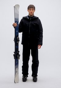 Μαύρο μπουφάν και παντελόνι για σκι, κρατώντας μπλε και άσπρα σκι με δεσίματα. Τα σκι έχουν γεωμετρικά σχέδια, με κωνικό σχήμα και λείο φινίρισμα.