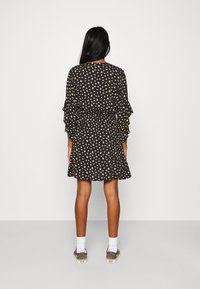 Schwarzes Kleid mit kleinem gelbem Blumendesign und Rüschensäumen, tailliert, ausgestellt am Saum, kombiniert mit weißen Socken und grünen Sneakers.