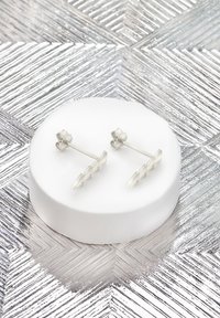 Orecchini a bottone in argento con design a zigzag, finitura lucida e chiusura a farfalla, esposti su un supporto circolare bianco.