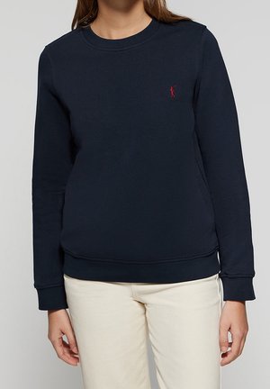 Person iført en mørkeblå langærmet sweatshirt med et lille rødt broderet logo, kombineret med lyse beige bukser.
