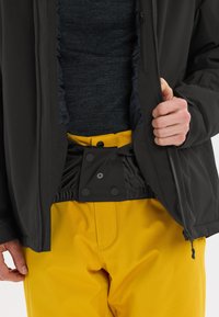 Schwarze Jacke, teilweise geöffnet, die ein dunkles strukturiertes Hemd darunter zeigt. Gelbe Hose mit Druckknopfverschluss und elastischem Bund sichtbar.