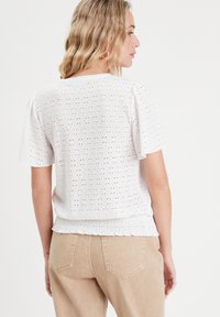 White, short-sleeved top with textured eyelet pattern, elasticated hem, and puffed sleeves. Worn with beige high-waisted pants.