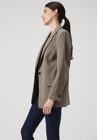 Hnědý kostkovaný blazer s klopou a jednou náprsní kapsou. Tkanina má strukturovaný povrch, s jemnými pruhy a černými knoflíky jako akcenty.