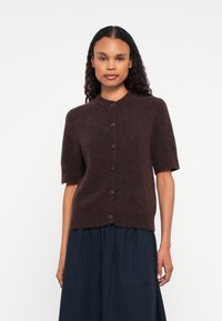 Mulher a usar um cardigan de mangas curtas em castanho escuro com botões e uma saia plissada azul escura diante de um fundo claro.