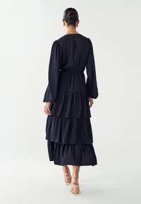 Morska dolga obleka z več ravnmi in dolgimi rokavi, z elastičnim pasom in padajočimi obroči. Gladek material z rahlim sijajem, v kombinaciji z čevlji s peto in gležnjem.