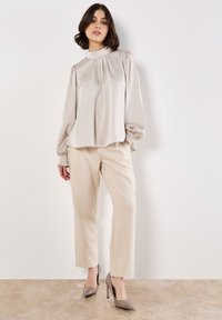Blusa color crema con righe verticali, collo alto e maniche a sbuffo, abbinata a pantaloni beige e tacchi alti taupe.