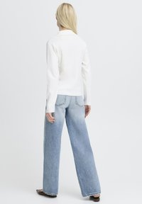 Femme blonde debout, vue de dos, portant un haut blanc à manches longues, un jean large bleu clair et des chaussures plates marron.