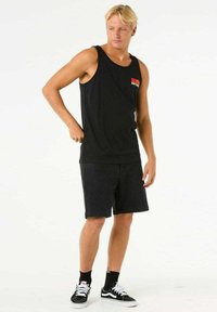 Schwarzes Tanktop mit einem roten und gelben Logo, kombiniert mit schwarzen Jeansshorts. Das Model trägt schwarze Socken und weiße Sneakers.
