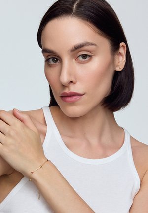 Złota bransoletka z okrągłymi akcentami koralików i małym czerwonym kamieniem, noszona na nadgarstku. Model ma na sobie top w neutralnym kolorze. Gładka tekstura skóry.