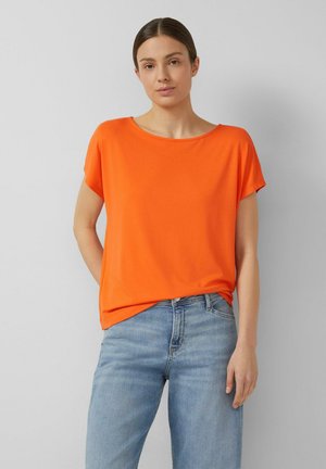 Młoda kobieta z ciemnymi włosami upiętymi w koka, ubrana w jaskrawą pomarańczową bluzkę z krótkim rękawem i jasnoniebieskie dżinsy, stojąca na szarym tle.
