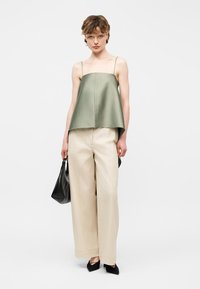 Γυναίκα με φαρδιά μπεζ παντελόνια, ελαφρώς φαρδιά ελιάς πράσινο αμάνικο τοπ, μαύρα μυτερά παπούτσια, κρατώντας μαύρη τσάντα, μπροστά σε απλό φόντο.