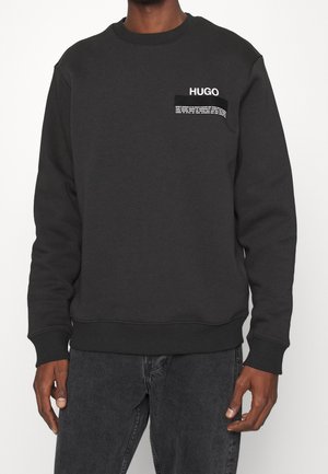 Sweat-shirt noir à manches longues avec le logo "HUGO" et un petit texte blanc sur la poitrine gauche, porté avec un jean gris foncé.