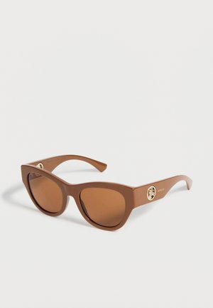 Brown cat-eye sunglasses with tinted lenses and gold circular emblem on the temple, placed on a white surface.