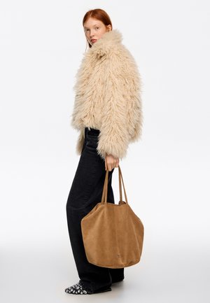 Bežs mākslīgās kažokādas žakete ar augstu apkakli, nēsāta pār melnām platām džinsa biksēm, rokās mīksta brūna zamša soma ar divām plecu siksnām.