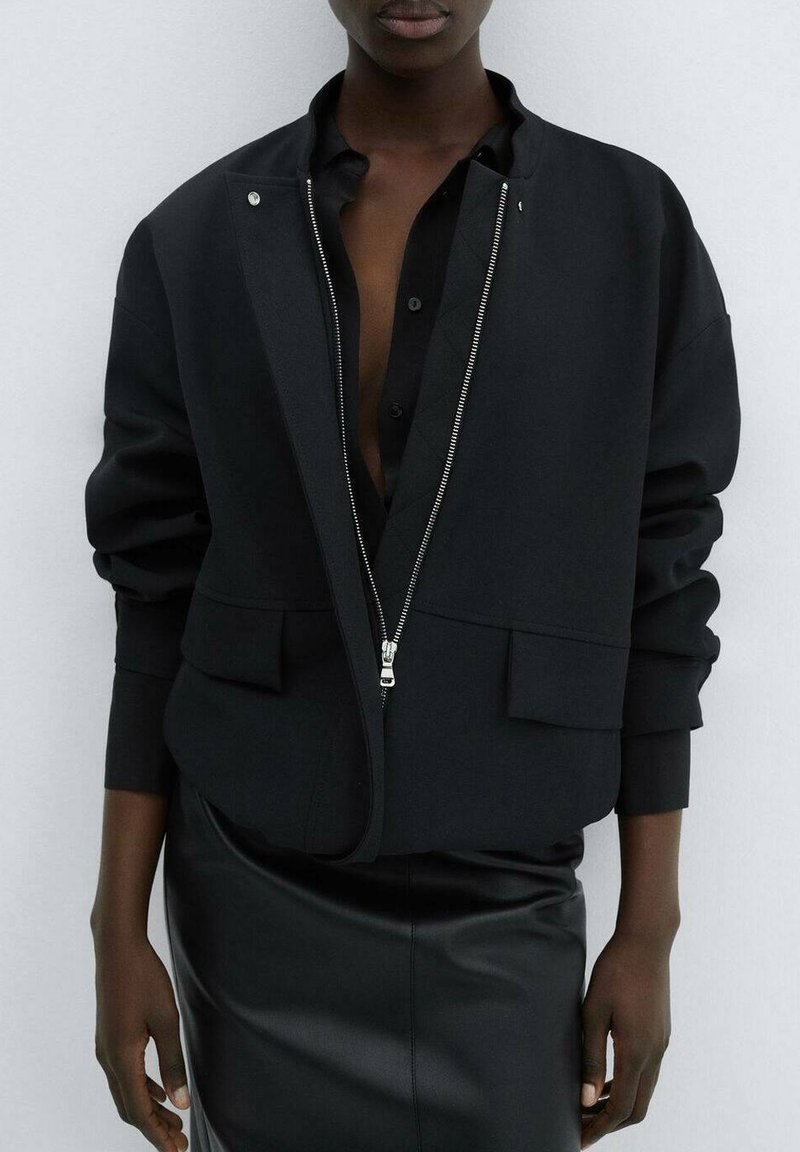 Personne portant une veste noire ouverte sur une chemise noire partiellement déboutonnée et une jupe en cuir noire, debout devant un fond clair.