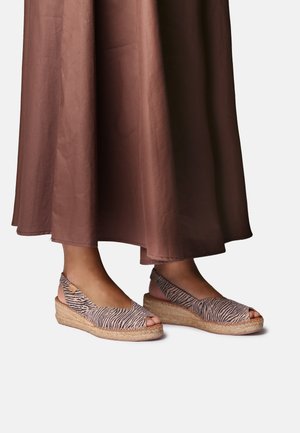 Voeten in open teen sleehakken met zebra-print bandjes, gecombineerd met een lange bruine rok tegen een effen lichte achtergrond.